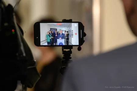 Goście uroczystości uruchomienia Oddziału Urazowo-Ortopedycznego w SP ZOZ MSWiA widoczni na ekranie telefonu komórkowego.