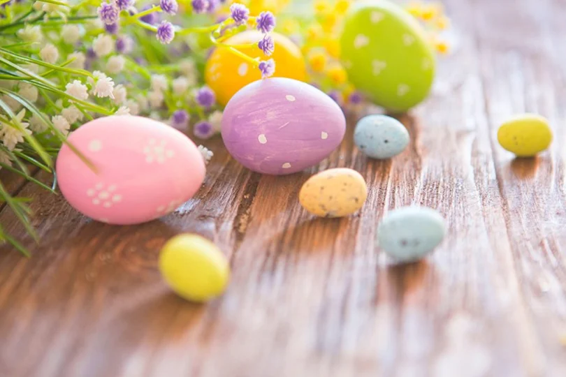 Wielkanocna grafika. Rozsypane na stole pastelowe pisanki oraz kwiaty.