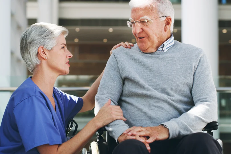 Zdjęcie przedstawia starszą osobę na wózku i opiekuna