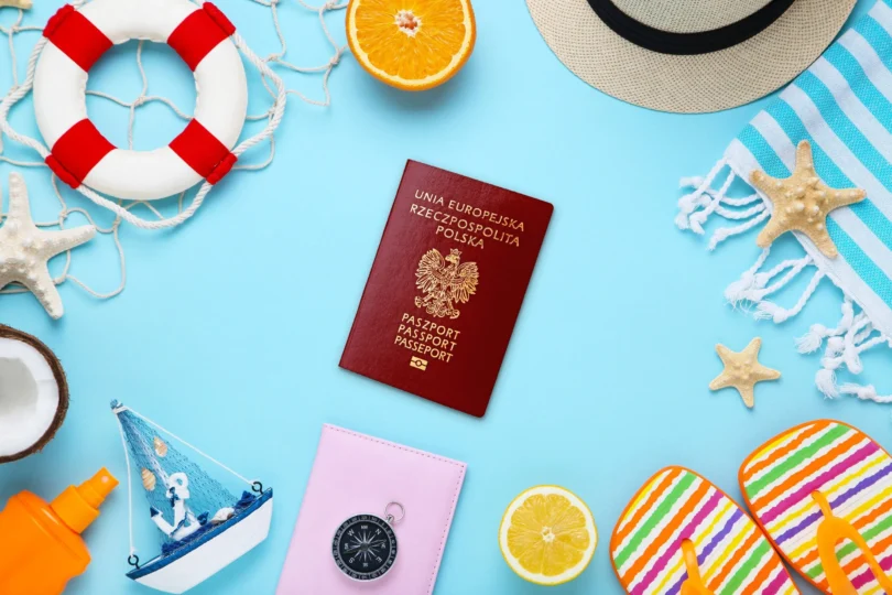 na niebieskim tle grafiki na środku widnieje paszport, a dookoła rzeczy kojarzące się z wakacjami: klapki plażowe, rozgwiazdy, ręcznik, kompas, koło ratunkowe, łódka, kapelusz, olejek do opalania i cząstki pomarańczy.